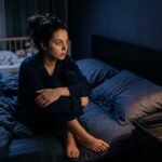 A tired new mother sitting awake in bed at night experiencing postpartum anxiety at night while her baby sleeps peacefully in a crib nearby